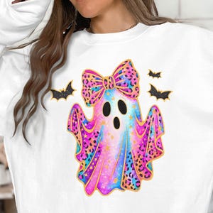 May include: White sweatshirt featuring a colorful ghost design with a bow, leopard print accents, and star details. The ghost has three black eye holes and is accompanied by three black bats with gold outlines. A festive Halloween-themed garment.