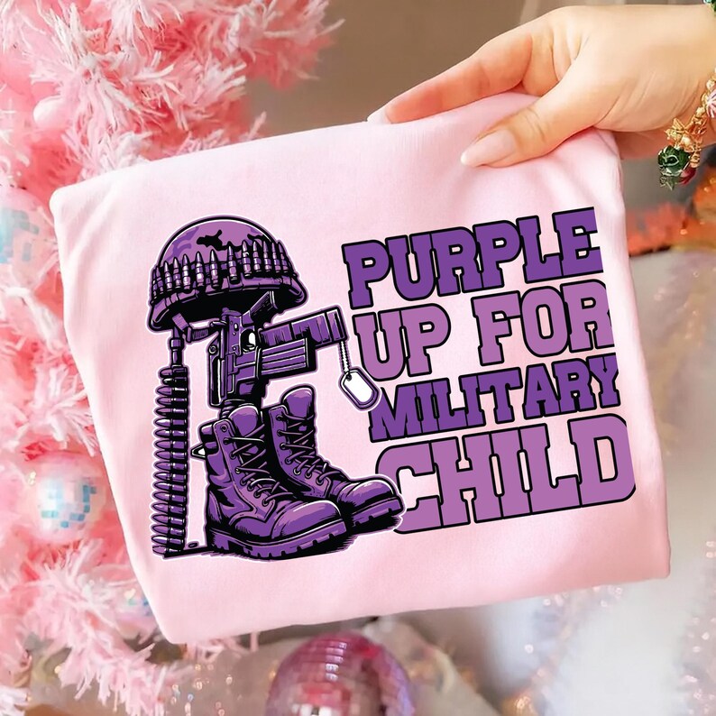 Puede incluir: Camiseta rosa claro con un gr&aacute;fico morado que representa un casco militar, un rifle, una chapa de identificaci&oacute;n y botas. El texto dice "PURPLE UP FOR MILITARY CHILD".