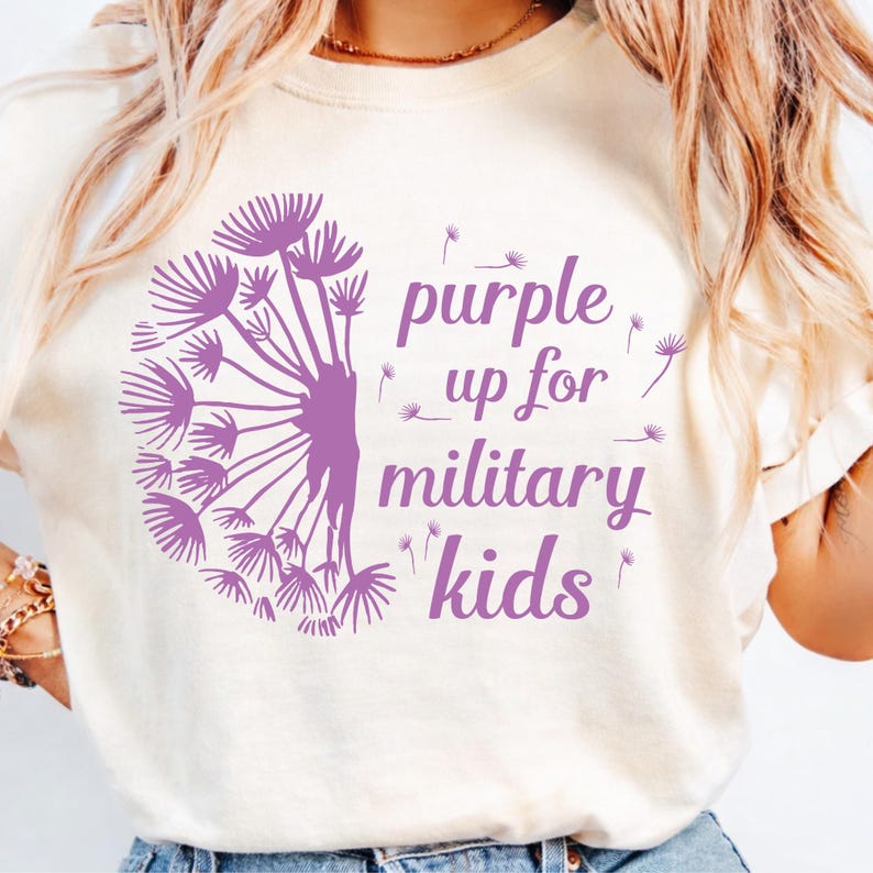 Puede incluir: Camiseta color crema con un gran dise&ntilde;o de diente de le&oacute;n morado. El texto "purple up for military kids" est&aacute; impreso en morado. Una prenda casual.