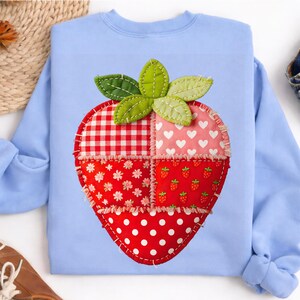 May include: A light blue sweatshirt featuring a large, appliqued strawberry design. The strawberry is composed of various red and pink patterned fabrics, including gingham, hearts, and polka dots, with green leaves at the top. The design is stitched onto the sweatshirt.