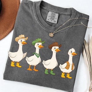 May include: Dark grey Comfort Colors t-shirt with a cartoon design of four white geese dressed in cowboy attire. The geese wear hats, boots, and vests. One goose has a green bandana. A necklace with a pendant is also visible.