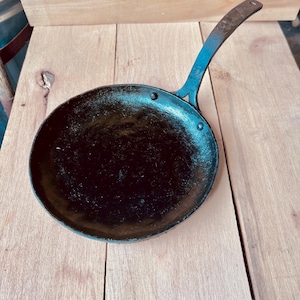 Op de afbeelding: Een zwarte, ronde gietijzeren koekenpan met een lange steel. De steel is met klinknagels aan de pan bevestigd. De pan staat op een lichtbruin houten oppervlak. De steel heeft een gat om op te hangen.