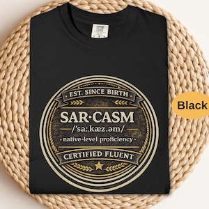 May include: Black t-shirt with a round, vintage-style graphic that reads "SAR-CASM" with the phonetic pronunciation and "CERTIFIED FLUENT." The graphic is in shades of gold and brown. The text "EST. SINCE BIRTH" is at the top.