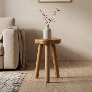 Könnte beinhalten: Ein kleiner, runder Hocker aus hellbraunem Holz mit drei zylindrischen Beinen. Auf dem Hocker steht eine matte, cremeweiße Keramikvase mit einem zarten Zweig, der kleine, hellrosa Blüten trägt, was ein schlichtes und funktionales Möbelstück ergibt.