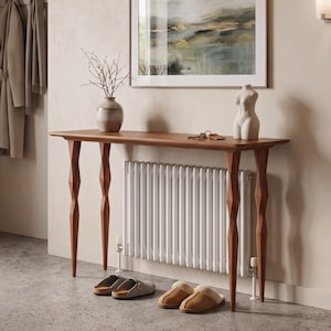 Peut inclure: Une console en bois dotée de quatre pieds aux formes distinctives et d'un plateau rectangulaire lisse. La table est placée contre un mur au-dessus d'un radiateur blanc, avec un vase en céramique, une petite sculpture et des clés sur le dessus.