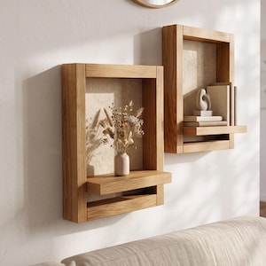 Pode incluir: Duas prateleiras de parede retangulares de madeira castanho-clara com acabamento em grão natural. Cada prateleira possui uma estrutura sólida e uma borda horizontal. Uma contém um vaso com flores secas, a outra livros e uma pequena escultura.
