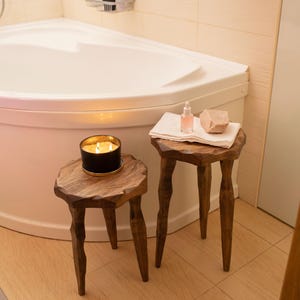 May include: Two dark brown wooden stools with three legs and uneven, natural-edge circular tops. One stool holds a black candle with a lit flame. The other stool holds a folded white towel, a small clear glass spray bottle, and a beige stone.
