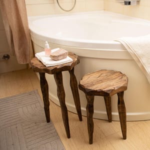 May include: Two small, rustic wooden stools with carved legs are in a bathroom. A white bathtub is in the background, with a folded white towel and a lit black candle. A small bottle and a stone sit on one of the stools.