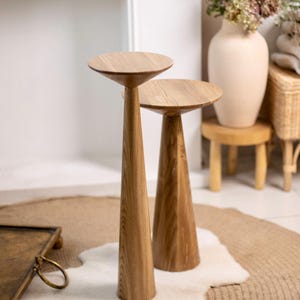 Puede incluir: Dos mesas auxiliares de madera con acabado natural. Las mesas tienen una parte superior redonda y una base cónica. Las mesas están sobre una alfombra con un acento de piel blanca. Un jarrón con flores, un taburete pequeño y un banco de almacenamiento tejido están en el fondo.