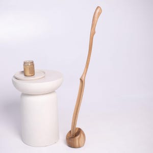 May include: A white, modern side table with a small gold-colored jar on top. A tall, wooden, curved sculpture stands beside the table. The sculpture has a rounded base and a slender, naturalistic form.