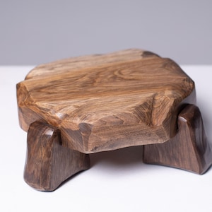 Może przedstawiać: Ręcznie wykonany drewniany stołek o unikalnym, kanciastym designie. Stołek ma naturalne wykończenie słojów drewna i mierzy około 30 cm długości, 25 cm szerokości i 20 cm wysokości. Nogi są solidne i zapewniają stabilną podstawę.