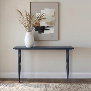 Puede incluir: Una consola azul marino con una tapa larga y rectangular y patas decorativas y anguladas. Un jarrón blanco con tallos de trigo seco se encuentra sobre la mesa. Una pintura abstracta cuelga de la pared detrás de la mesa.