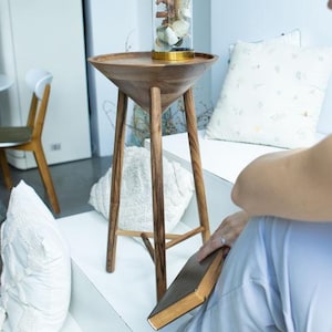 May include: A wooden side table with a conical top and three angled legs. A glass cloche with a gold base sits on the table, displaying a decorative arrangement. The table is in a bright, modern interior setting.