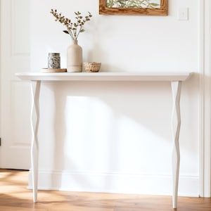 May include: A white console table with a rectangular top and decorative, geometric legs. The table is in a bright room with a vase, small basket, and other decorative items on top. The table is approximately 48 inches long.