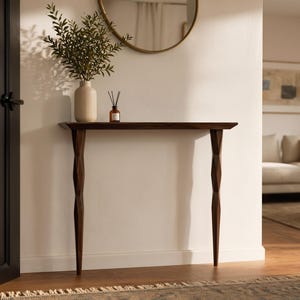 Puede incluir: Una consola de madera marrón oscuro con una parte superior estrecha y patas talladas intrincadamente. Un jarrón con vegetación, un difusor de caña y un espejo redondo están en la pared detrás de la mesa. La mesa es una adición elegante a cualquier entrada o sala de estar.
