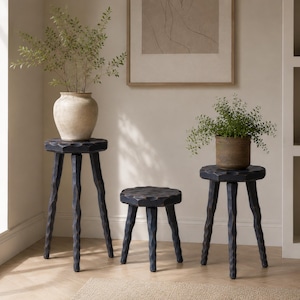 May include: Three dark charcoal-colored wooden stools with textured, faceted surfaces and three legs each. The stools vary in height, with two taller stools supporting potted plants and one shorter stool positioned in the center of the group.