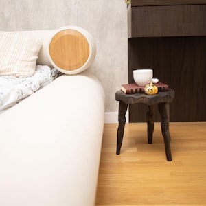 May include: A white bed with a round headboard featuring a wooden accent. A small, dark wooden side table holds a book, a white cup, and a decorative pumpkin. The room has light-colored walls and wooden flooring.