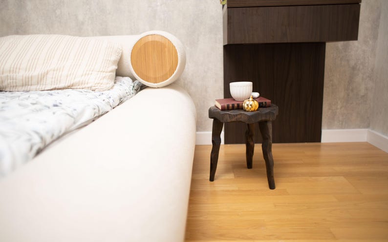 May include: A white bed with a round headboard featuring a wooden accent. A small, dark wooden side table holds a book, a white cup, and a decorative pumpkin. The room has light-colored walls and wooden flooring.
