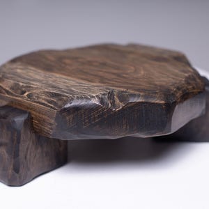 Puede incluir: Bandeja de madera tallada a mano de color marr&oacute;n oscuro o soporte de exhibici&oacute;n. La bandeja tiene una forma natural y org&aacute;nica con una textura &aacute;spera y se apoya en tres patas robustas y te&ntilde;idas de oscuro. La veta de la madera es visible.