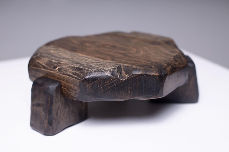 Puede incluir: Bandeja de madera tallada a mano de color marr&oacute;n oscuro o soporte de exhibici&oacute;n. La bandeja tiene una forma natural y org&aacute;nica con una textura &aacute;spera y se apoya en tres patas robustas y te&ntilde;idas de oscuro. La veta de la madera es visible.