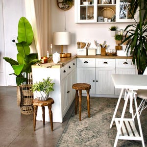 May include: A bright, airy kitchen with white cabinets, a wooden countertop, and a white table. The room is decorated with plants, including a large potted plant with broad green leaves. Two wooden stools and a woven basket add to the natural aesthetic.