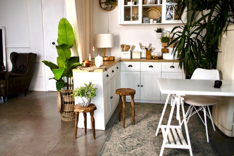 May include: A bright, airy kitchen with white cabinets, a wooden countertop, and a white table. The room is decorated with plants, including a large potted plant with broad green leaves. Two wooden stools and a woven basket add to the natural aesthetic.