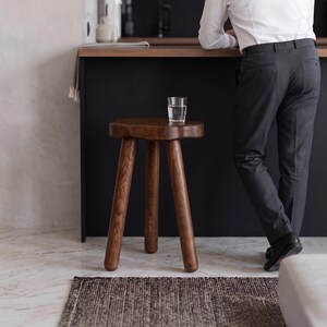 May include: A three-legged wooden stool with a dark brown finish and a rounded, organic-shaped seat. The stool features a smooth, polished surface showing the natural wood grain, supported by three sturdy, cylindrical legs angled outward.