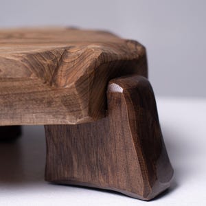 Puede incluir: Tabla de servir de madera marr&oacute;n oscuro con borde natural. Se apoya en dos patas anguladas, creando una base estable. La veta de la madera es visible. La tabla mide aproximadamente 30 cm de largo.