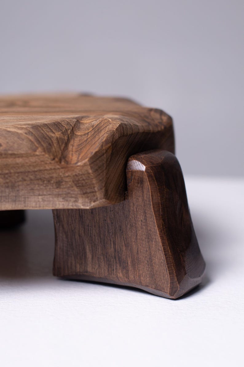 Puede incluir: Tabla de servir de madera marr&oacute;n oscuro con borde natural. Se apoya en dos patas anguladas, creando una base estable. La veta de la madera es visible. La tabla mide aproximadamente 30 cm de largo.