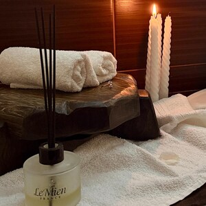 Puede incluir: Una escena de spa con toallas blancas enrolladas, velas encendidas y un difusor de ca&ntilde;a. La botella del difusor, etiquetada "Le Mien France", est&aacute; frente a una plataforma de madera oscura. Las velas son blancas y retorcidas.
