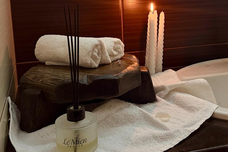 Puede incluir: Una escena de spa con toallas blancas enrolladas, velas encendidas y un difusor de ca&ntilde;a. La botella del difusor, etiquetada "Le Mien France", est&aacute; frente a una plataforma de madera oscura. Las velas son blancas y retorcidas.