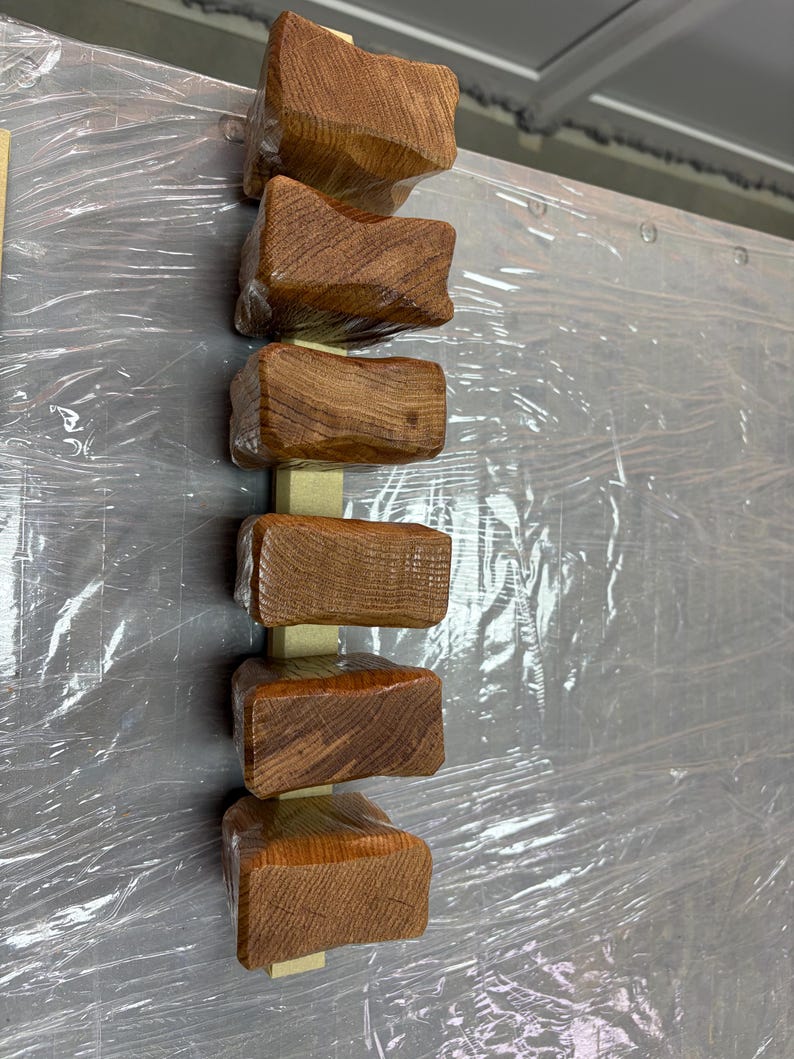 Puede incluir: Disposici&oacute;n vertical de seis bloques de madera tallada, cada uno con una superficie texturizada &uacute;nica y un tono marr&oacute;n c&aacute;lido. Los bloques est&aacute;n unidos a un soporte de madera de color claro, creando una est&eacute;tica r&uacute;stica y artesanal.
