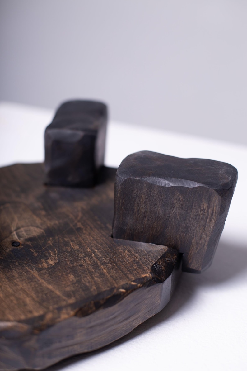 Puede incluir: Una bandeja de madera tallada a mano, de color marr&oacute;n oscuro, con tres patas cuadradas en forma de bloques. La bandeja tiene una veta de madera natural y una est&eacute;tica r&uacute;stica y org&aacute;nica. Las patas est&aacute;n unidas a la base de la bandeja.