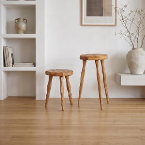 May include: Two wooden stools with round, textured tops and three wavy, carved legs. The stools are crafted from light-toned wood with a natural grain finish. One stool is shorter than the other, and both feature a distinct, organic, hand-carved appearance.