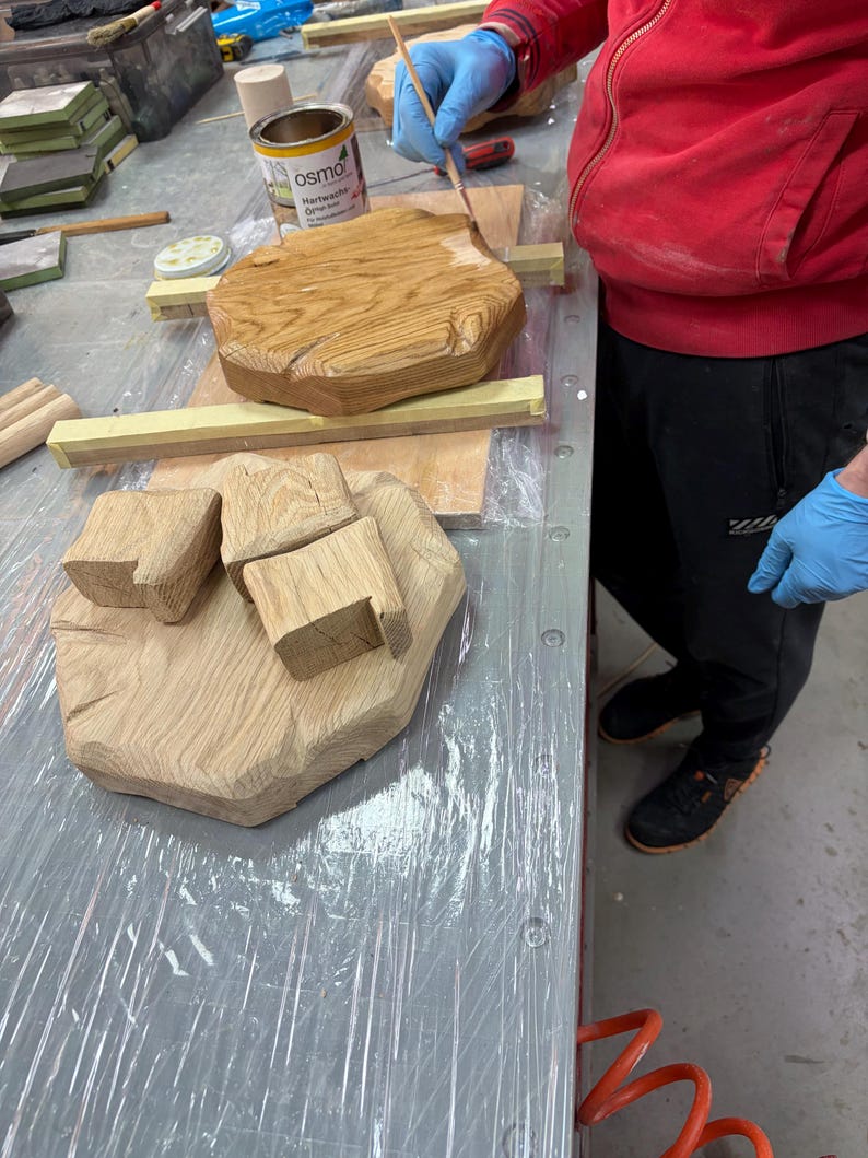 Puede incluir: Una persona aplica un acabado de madera a un objeto de madera de color claro y forma irregular. El objeto est&aacute; sobre una superficie de trabajo con otras piezas de madera. Se ve una lata de Osmo Hartwachs-&Ouml;l.
