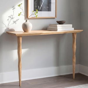 May include: A light oak console table with a rectangular top and two carved legs. The table features a vase with greenery, a stack of books, and a small bowl. The table is positioned against a light grey wall. The table's dimensions are in cm.