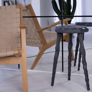 May include: A modern interior scene featuring a woven leather chair and a wooden armchair. Two dark gray, three-legged stools with carved legs are in the foreground. The stools have a hexagonal seat. A large mirror reflects the scene.