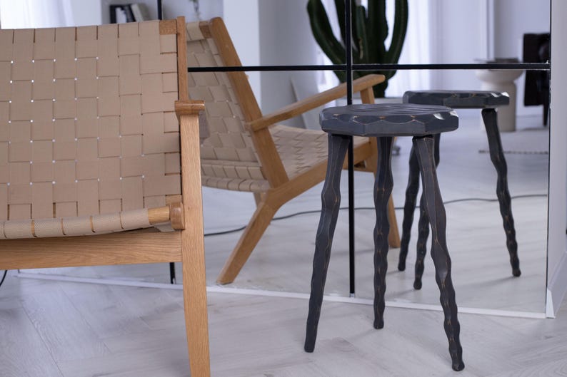 May include: A modern interior scene featuring a woven leather chair and a wooden armchair. Two dark gray, three-legged stools with carved legs are in the foreground. The stools have a hexagonal seat. A large mirror reflects the scene.