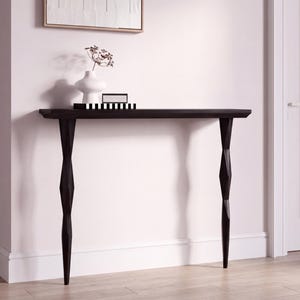 Könnte beinhalten: Eine dunkelbraune Konsole mit rechteckiger Platte und zwei dekorativen Beinen. Die Beine haben ein einzigartiges, verdrehtes Design. Eine weiße Vase mit Trockenblumen und eine dekorative Box stehen auf dem Tisch.