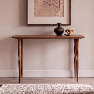 May include: A rectangular wooden console table with four slender, uniquely shaped legs. The table surface holds a dark brown ceramic vase and a decorative piece of natural coral. The wood has a warm, medium-brown tone and a smooth finish.