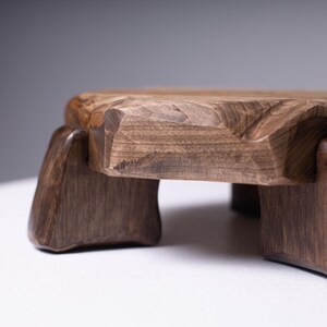 Puede incluir: Un taburete de madera hecho a mano con un acabado marr&oacute;n oscuro. El taburete tiene una parte superior cuadrada con bordes tallados y cuatro patas robustas. La veta de la madera es visible, lo que se suma a su est&eacute;tica natural. Este peque&ntilde;o taburete decorativo es adecuado para varios usos.