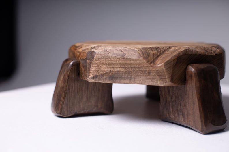 Puede incluir: Un taburete de madera hecho a mano con un acabado marr&oacute;n oscuro. El taburete tiene una parte superior cuadrada con bordes tallados y cuatro patas robustas. La veta de la madera es visible, lo que se suma a su est&eacute;tica natural. Este peque&ntilde;o taburete decorativo es adecuado para varios usos.
