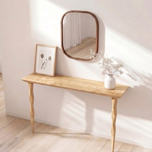 May include: A light wood console table with a framed print and a brown-framed mirror. A white vase with white flowers sits on the table. The table has a rectangular top and carved legs. The mirror is oval-shaped.