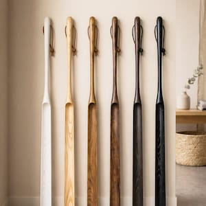 May include: Six wooden shoe horns in varying natural wood finishes are displayed against a neutral wall. The colors range from white to black, with leather loops at the top. The text "6 Natural wood finishes" is at the top.