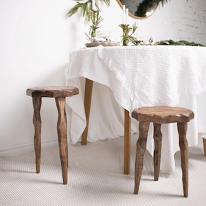 May include: Two handcrafted wooden stools with unique, organic-shaped legs and tops. The stools are made of a warm-toned wood and are placed on a light-colored rug. A round table with a white tablecloth is in the background.