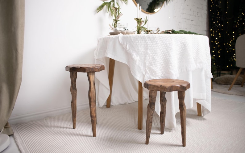 May include: Two handcrafted wooden stools with unique, organic-shaped legs and tops. The stools are made of a warm-toned wood and are placed on a light-colored rug. A round table with a white tablecloth is in the background.