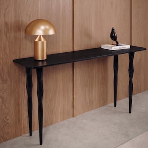 Può includere: Un tavolo console nero con quattro gambe affusolate e sfaccettate. Sul tavolo sono poggiati una lampada dorata a forma di fungo, un libro bianco intitolato Bauhaus e una piccola statuetta di uccello nero. Il tavolo ha un piano rettangolare.