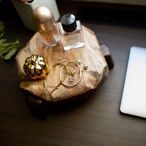 Puede incluir: Una bandeja decorativa de madera con un frasco de perfume dorado, un frasco de perfume peque&ntilde;o, una calabaza dorada y joyas doradas. Un port&aacute;til plateado est&aacute; a la derecha. Una planta est&aacute; en el fondo.