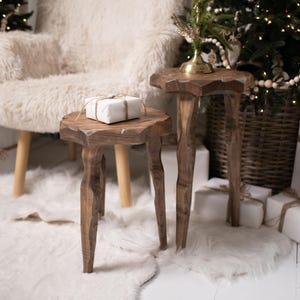 May include: Two rustic wooden side tables with unique, organic shapes. One table has a wrapped gift on top. The tables are set on a white rug, with a fluffy armchair and Christmas trees in the background. The scene evokes a cozy, festive atmosphere.