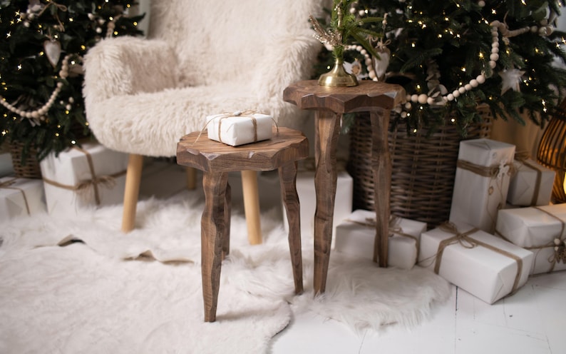May include: Two rustic wooden side tables with unique, organic shapes. One table has a wrapped gift on top. The tables are set on a white rug, with a fluffy armchair and Christmas trees in the background. The scene evokes a cozy, festive atmosphere.